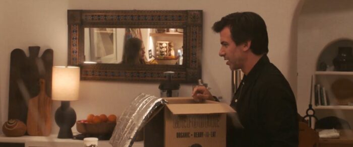 Asher talks to Whitney, who is seen reflected in a mirror in their home