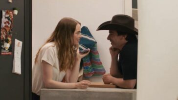 Whitney and Asher look at each other while leaning on a counter in The Curse S1E4. She's holding a phone. He's wearing a cowboy hat