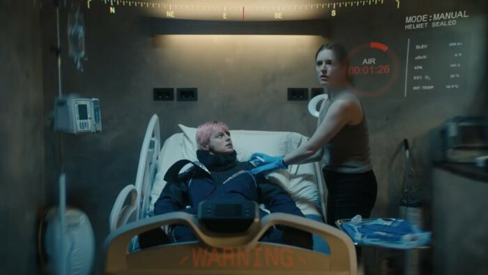 Eva stands over Darby on a medical bed, viewed through the heads up display include a helmet