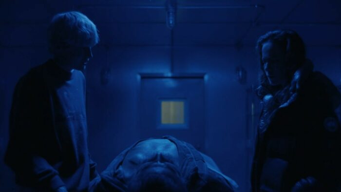 A Murder at the End of the World S1E4 - Darby and Sian stand over Rohan's body in the kitchen freezer, blue lit