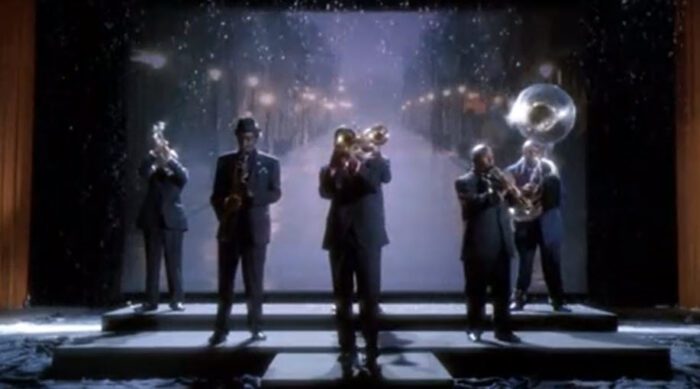 A brass quintet stands on a dramatically lit stage, with a screen behind them showing photos of New Orleans