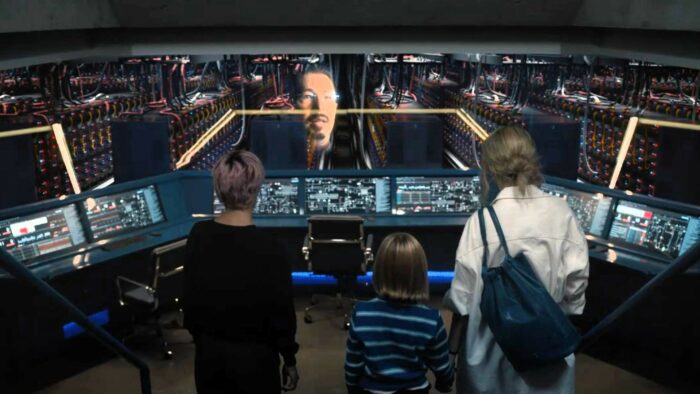 Darby, Zoomer and Lee stand in the control room looking at a projection of Ray's face, overlaid on the view of a vast server room