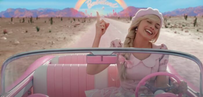Margot Robbie as Barbie points a finger into the air as she drives her pink car