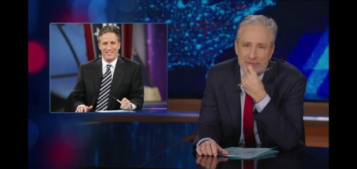Jon Stewart sits at the Daily Show desk, with a picture of a younger version of himself in the corner of the screen