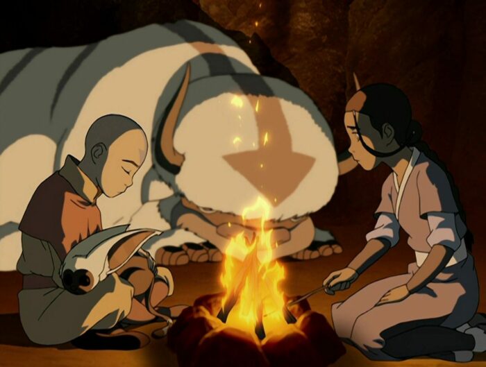 Aang with Momo sleeping in his lap sits in front of a fire by Katara with Appa in the background