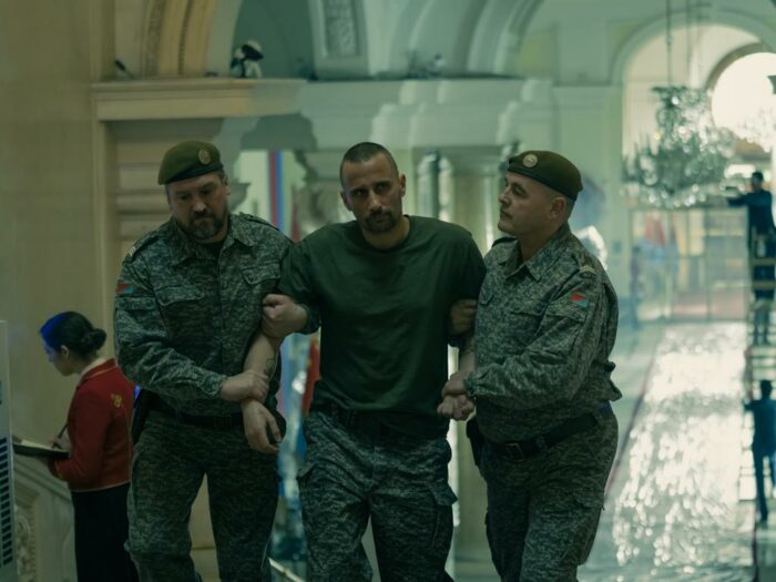 Zubak is held by two soldiers as they walk him into the palace, with other workers and plastic covered walls in the background