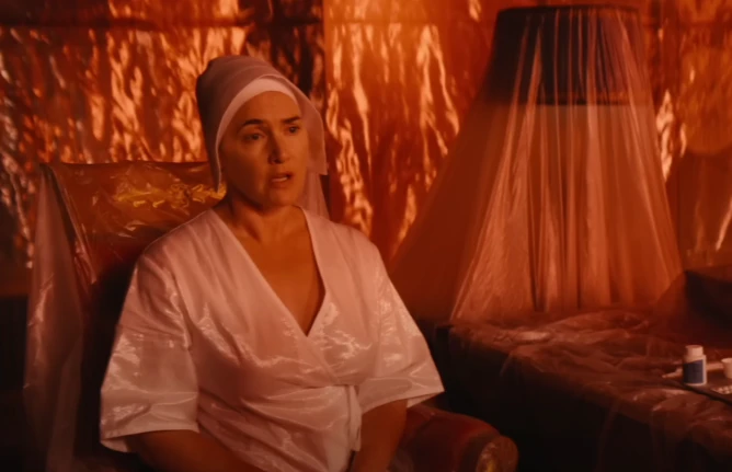 Kate Winslet in a white gown and hospital cap in a red tented room covered in plastic tarps