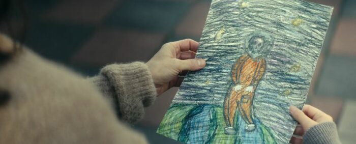 A hand holding a drawing of a dead person in space wearing orange