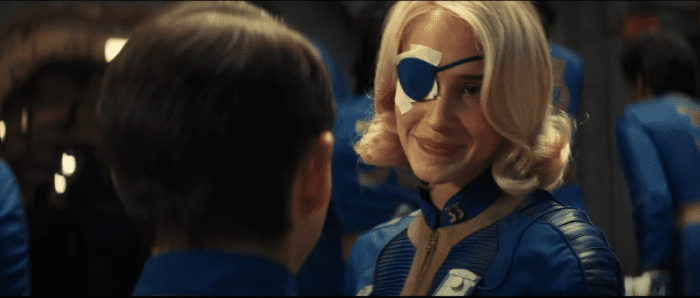 A woman in a blue jumpsuit with long blonde hair wearing an eye patch smiles at a young man facing her 