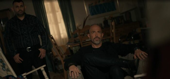 Stallings sits in a chair in Teresa's apartment, flanked by a goon